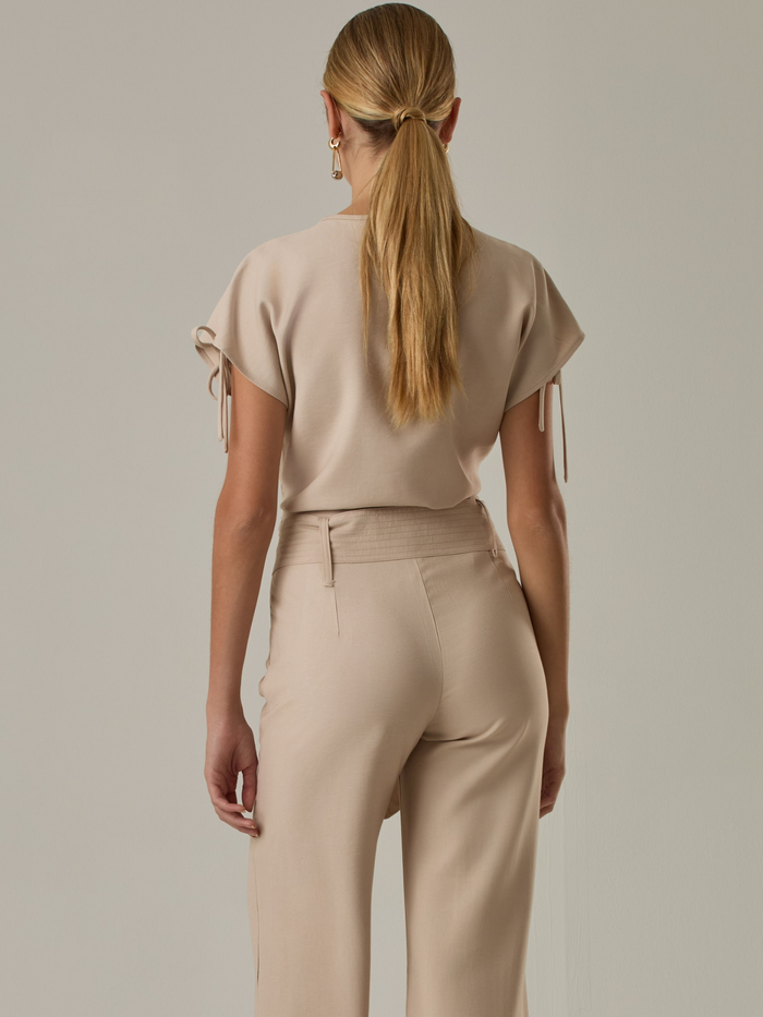 Blusa con  diseño elegante y minimalista. Presenta un sutil escote en forma de lágrima en la parte frontal, mangas cortas con lazos ajustables en los laterales que le aportan un toque delicado y femenino. Su tejido ligero y de textura suave le otorga comodidad y sofisticación.
