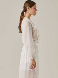 Blusa de manga larga confeccionada en un tejido ligero de textura sutil. Presenta un escote en "V" adornado con un amarre de tiras cruzadas, que añade un toque delicado al diseño. El frente está decorado con volantes que caen en capas suaves,
