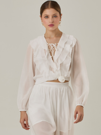 Blusa de manga larga confeccionada en un tejido ligero de textura sutil. Presenta un escote en "V" adornado con un amarre de tiras cruzadas, que añade un toque delicado al diseño. El frente está decorado con volantes que caen en capas suaves, aportando movimiento y un aire romántico. Las mangas tienen un ligero fruncido en los puños, creando un acabado abullonado discreto.

Detalles:

Color: Blanco Perla

Cuidado: Lavar a mano con jabón suave, por separado. No dejar en remojo. Planchar a temperatura alta.

