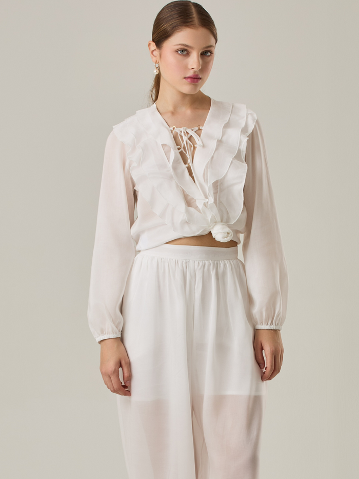 Blusa de manga larga confeccionada en un tejido ligero de textura sutil. Presenta un escote en "V" adornado con un amarre de tiras cruzadas, que añade un toque delicado al diseño. El frente está decorado con volantes que caen en capas suaves, aportando movimiento y un aire romántico. Las mangas tienen un ligero fruncido en los puños, creando un acabado abullonado discreto.

Detalles:

Color: Blanco Perla

Cuidado: Lavar a mano con jabón suave, por separado. No dejar en remojo. Planchar a temperatura alta.

