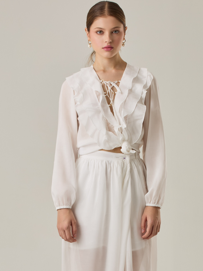 Blusa de manga larga confeccionada en un tejido ligero de textura sutil. Presenta un escote en "V" adornado con un amarre de tiras cruzadas, que añade un toque delicado al diseño. El frente está decorado con volantes que caen en capas suaves, aportando movimiento y un aire romántico. Las mangas tienen un ligero fruncido en los puños, creando un acabado abullonado discreto.

Detalles:

Color: Blanco Perla

Cuidado: Lavar a mano con jabón suave, por separado. No dejar en remojo.