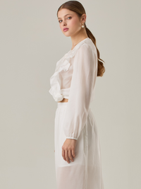 Blusa de manga larga confeccionada en un tejido ligero de textura sutil. Presenta un escote en "V" adornado con un amarre de tiras cruzadas, que añade un toque delicado al diseño. El frente está decorado con volantes que caen en capas suaves,