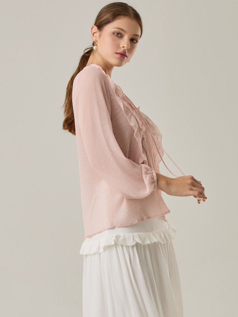Blusa de manga larga confeccionada en un tejido ligero de textura sutil. Presenta un escote en "V" adornado con un amarre de tiras cruzadas, que añade un toque delicado al diseño. El frente está decorado con volantes que caen en capas suaves, aportando movimiento y un aire romántico. Las mangas tienen un ligero fruncido en los puños, creando un acabado abullonado discreto.

Detalles:

Color: Rosa