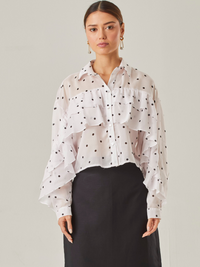 Esta blusa vaporosa de silueta relajada combina un estampado clásico de lunares con detalles delicados. Tiene cuello camisero, abotonadura frontal y volantes diagonales que recorren tanto el frente como las mangas, aportando movimiento y volumen. Su acabado semitransparente realza la ligereza de la prenda, perfecta para looks sofisticados con un toque divertido