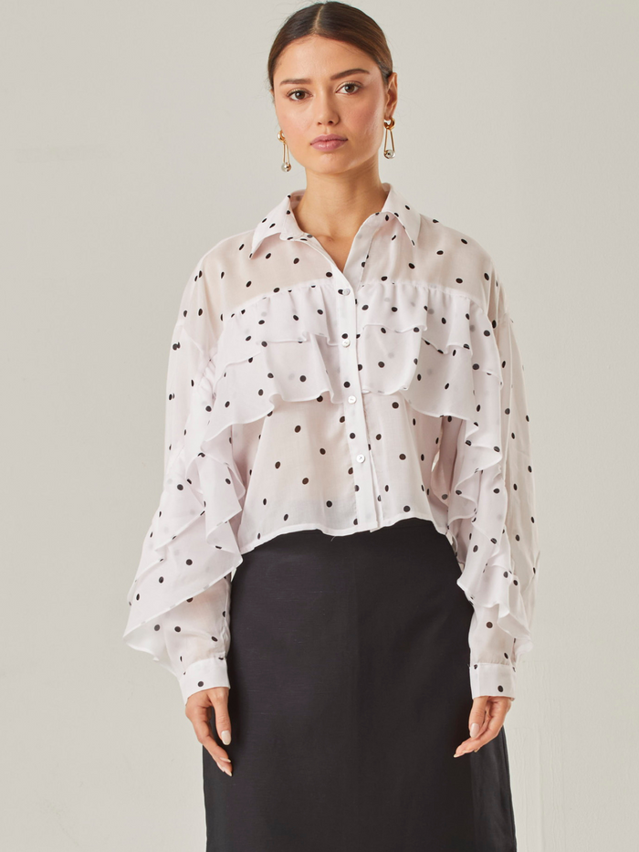Esta blusa vaporosa de silueta relajada combina un estampado clásico de lunares con detalles delicados. Tiene cuello camisero, abotonadura frontal y volantes diagonales que recorren tanto el frente como las mangas, aportando movimiento y volumen. Su acabado semitransparente realza la ligereza de la prenda, perfecta para looks sofisticados con un toque divertido