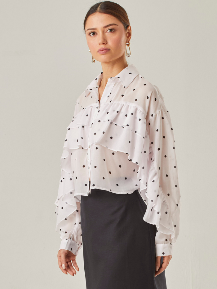Esta blusa vaporosa de silueta relajada combina un estampado clásico de lunares con detalles delicados. Tiene cuello camisero, abotonadura frontal y volantes diagonales que recorren tanto el frente como las mangas, aportando movimiento y volumen. Su acabado semitransparente realza la ligereza de la prenda, perfecta para looks sofisticados con un toque divertido