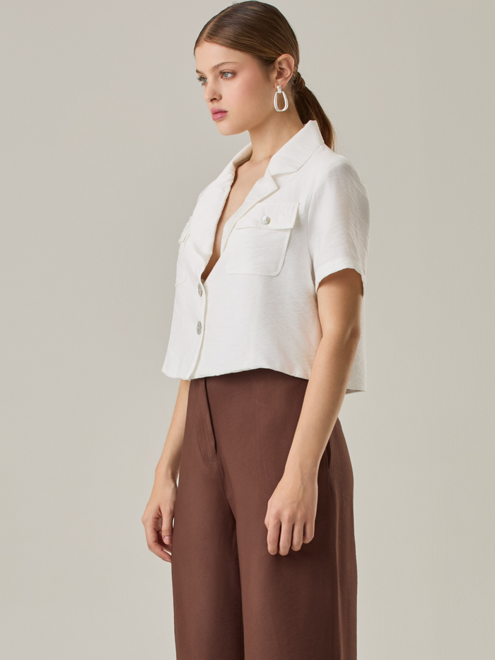 Esta blusa con cuello tipo sastre y mangas cortas aporta un aire fresco y contemporáneo. Los bolsillos con solapa en el frente agregan un toque funcional sin perder elegancia. Su corte ligeramente recortado la hace ideal para combinar con pantalones o faldas de tiro alto, creando un look moderno y pulido.