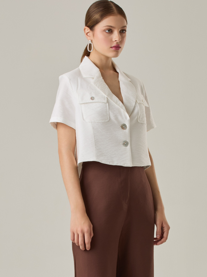 Esta blusa con cuello tipo sastre y mangas cortas aporta un aire fresco y contemporáneo. Los bolsillos con solapa en el frente agregan un toque funcional sin perder elegancia. Su corte ligeramente recortado la hace ideal para combinar con pantalones o faldas de tiro alto, creando un look moderno y pulido.