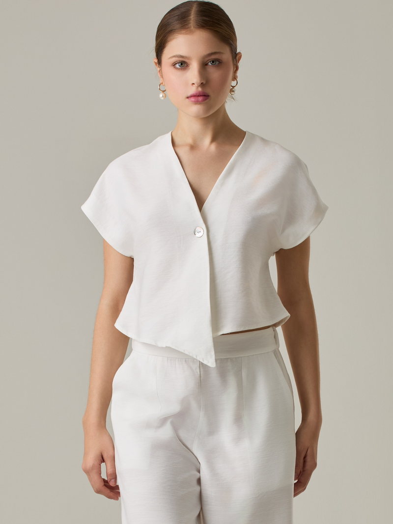 Blusa corta con silueta asimétrica y escote en V profundo. El diseño se cruza en el frente y se asegura con un botón al centro, generando una caída fluida y ligera. Las mangas cortas tipo capa aportan un aire moderno y relajado. Una prenda que combina líneas limpias con un toque contemporáneo.

Detalles:

Color: Blanco Perla

Material: 92% Rayón 8% Poliéster 