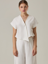 Blusa corta con silueta asimétrica y escote en V profundo. El diseño se cruza en el frente y se asegura con un botón al centro, generando una caída fluida y ligera. Las mangas cortas tipo capa aportan un aire moderno y relajado. Una prenda que combina líneas limpias con un toque contemporáneo.

Detalles:

Color: Blanco Perla

Material: 92% Rayón 8% Poliéster 