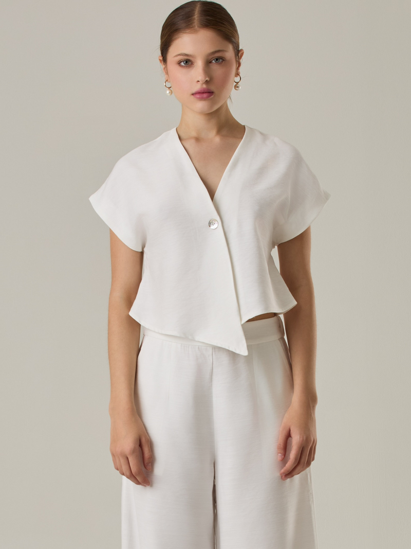 Blusa corta con silueta asimétrica y escote en V profundo. El diseño se cruza en el frente y se asegura con un botón al centro, generando una caída fluida y ligera. Las mangas cortas tipo capa aportan un aire moderno y relajado. Una prenda que combina líneas limpias con un toque contemporáneo.

Detalles:

Color: Blanco Perla

Material: 92% Rayón 8% Poliéster 