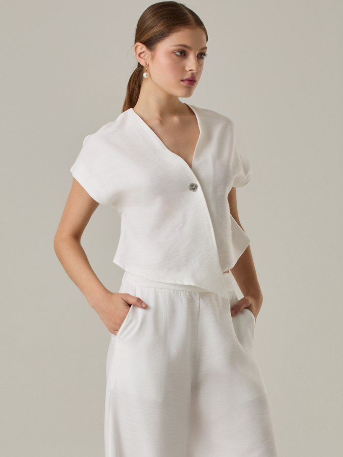 Blusa corta con silueta asimétrica y escote en V profundo. El diseño se cruza en el frente y se asegura con un botón al centro, generando una caída fluida y ligera. Las mangas cortas tipo capa aportan un aire moderno y relajado. Una prenda que combina líneas limpias con un toque contemporáneo.

Detalles:

Color: Blanco Perla