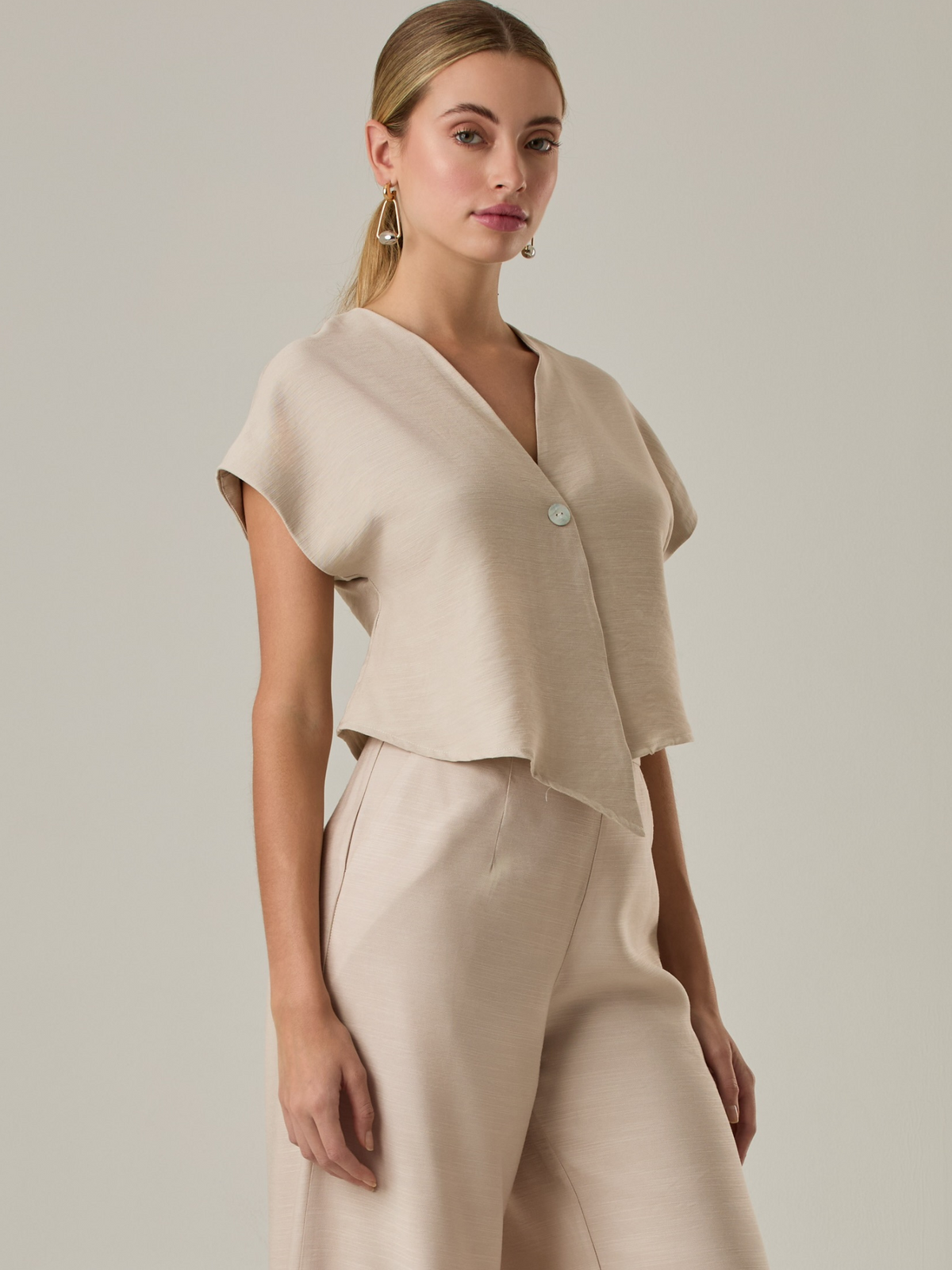 Blusa corta con silueta asimétrica y escote en V profundo. El diseño se cruza en el frente y se asegura con un botón al centro, generando una caída fluida y ligera. Las mangas cortas tipo capa aportan un aire moderno y relajado. Una prenda que combina líneas limpias con un toque contemporáneo.