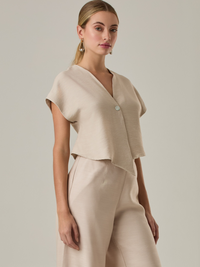 Blusa corta con silueta asimétrica y escote en V profundo. El diseño se cruza en el frente y se asegura con un botón al centro, generando una caída fluida y ligera. Las mangas cortas tipo capa aportan un aire moderno y relajado. Una prenda que combina líneas limpias con un toque contemporáneo.
