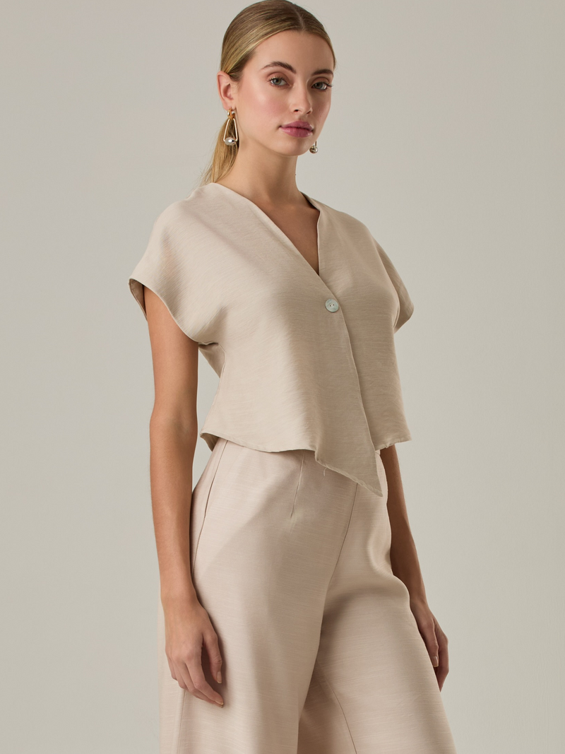 Blusa corta con silueta asimétrica y escote en V profundo. El diseño se cruza en el frente y se asegura con un botón al centro, generando una caída fluida y ligera. Las mangas cortas tipo capa aportan un aire moderno y relajado. Una prenda que combina líneas limpias con un toque contemporáneo.