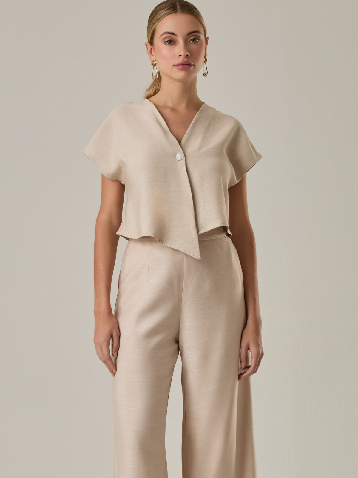 Blusa corta con silueta asimétrica y escote en V profundo. El diseño se cruza en el frente y se asegura con un botón al centro, generando una caída fluida y ligera. Las mangas cortas tipo capa aportan un aire moderno y relajado. Una prenda que combina líneas limpias con un toque contemporáneo.