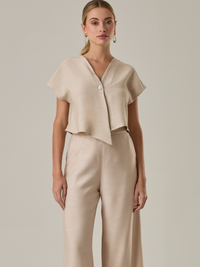 Blusa corta con silueta asimétrica y escote en V profundo. El diseño se cruza en el frente y se asegura con un botón al centro, generando una caída fluida y ligera. Las mangas cortas tipo capa aportan un aire moderno y relajado. Una prenda que combina líneas limpias con un toque contemporáneo.
