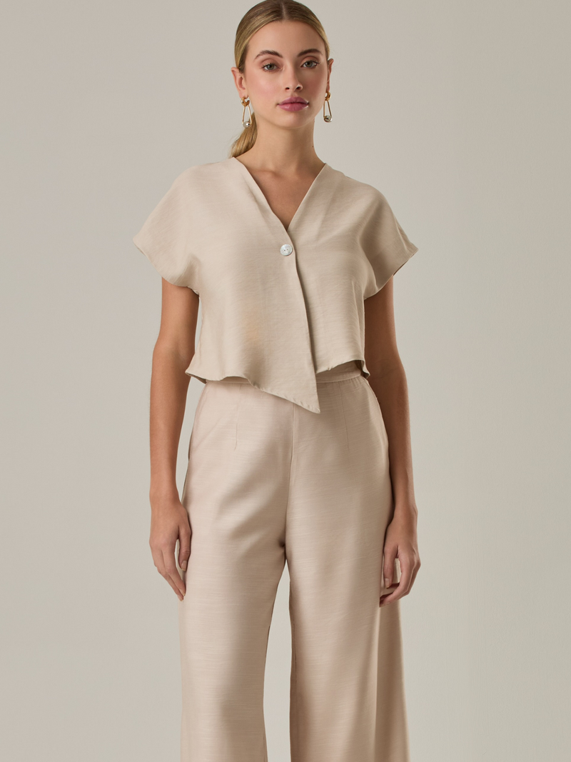 Blusa corta con silueta asimétrica y escote en V profundo. El diseño se cruza en el frente y se asegura con un botón al centro, generando una caída fluida y ligera. Las mangas cortas tipo capa aportan un aire moderno y relajado. Una prenda que combina líneas limpias con un toque contemporáneo.