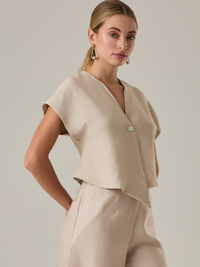 Blusa corta con silueta asimétrica y escote en V profundo. El diseño se cruza en el frente y se asegura con un botón al centro, generando una caída fluida y ligera. Las mangas cortas tipo capa aportan un aire moderno y relajado. Una prenda que combina líneas limpias con un toque contemporáneo.
