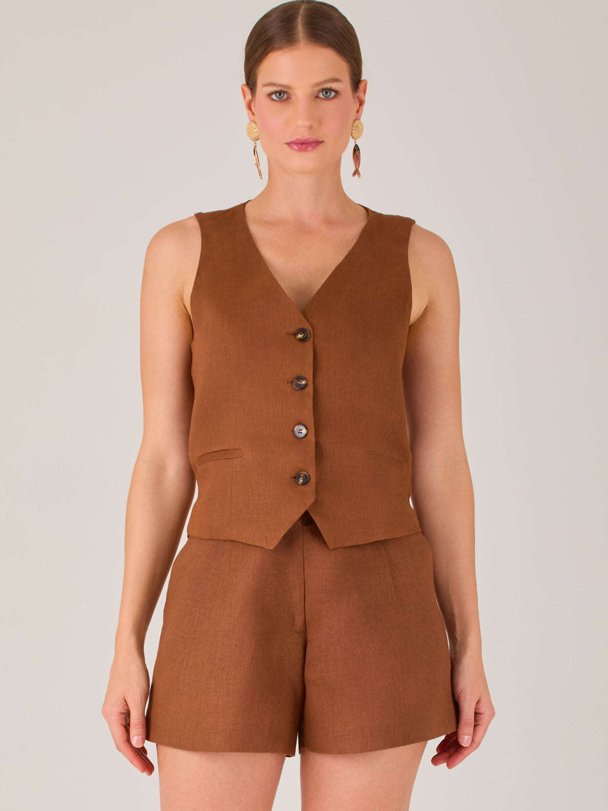 Esta blusa tipo chaleco es ideal para climas cálidos o para crear looks sofisticados con un aire casual. Presenta un corte recto ligeramente entallado que acentúa la silueta de forma sutil, y un escote en “V” que estiliza el cuello. Cuenta con una botonadura frontal compuesta por cinco botones oscuros que contrastan elegantemente con el tono neutro de la prenda. También incorpora bolsillos falsos decorativos a ambos lados del frente, que añaden un detalle sutil al diseño minimalista. Esta pieza es versátil 