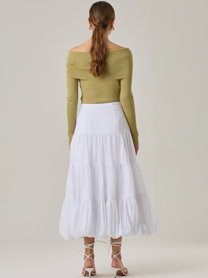 Esta es una falda blanca de largo midi con un diseño en capas que crea un efecto abombado en la parte inferior. Está hecha en una tela liviana y con caída. Tiene un estilo romántico y femenino, ideal para combinar con blusas ajustadas o crop tops. En la imagen se lleva con un suéter off-shoulder que le da un look sofisticado.