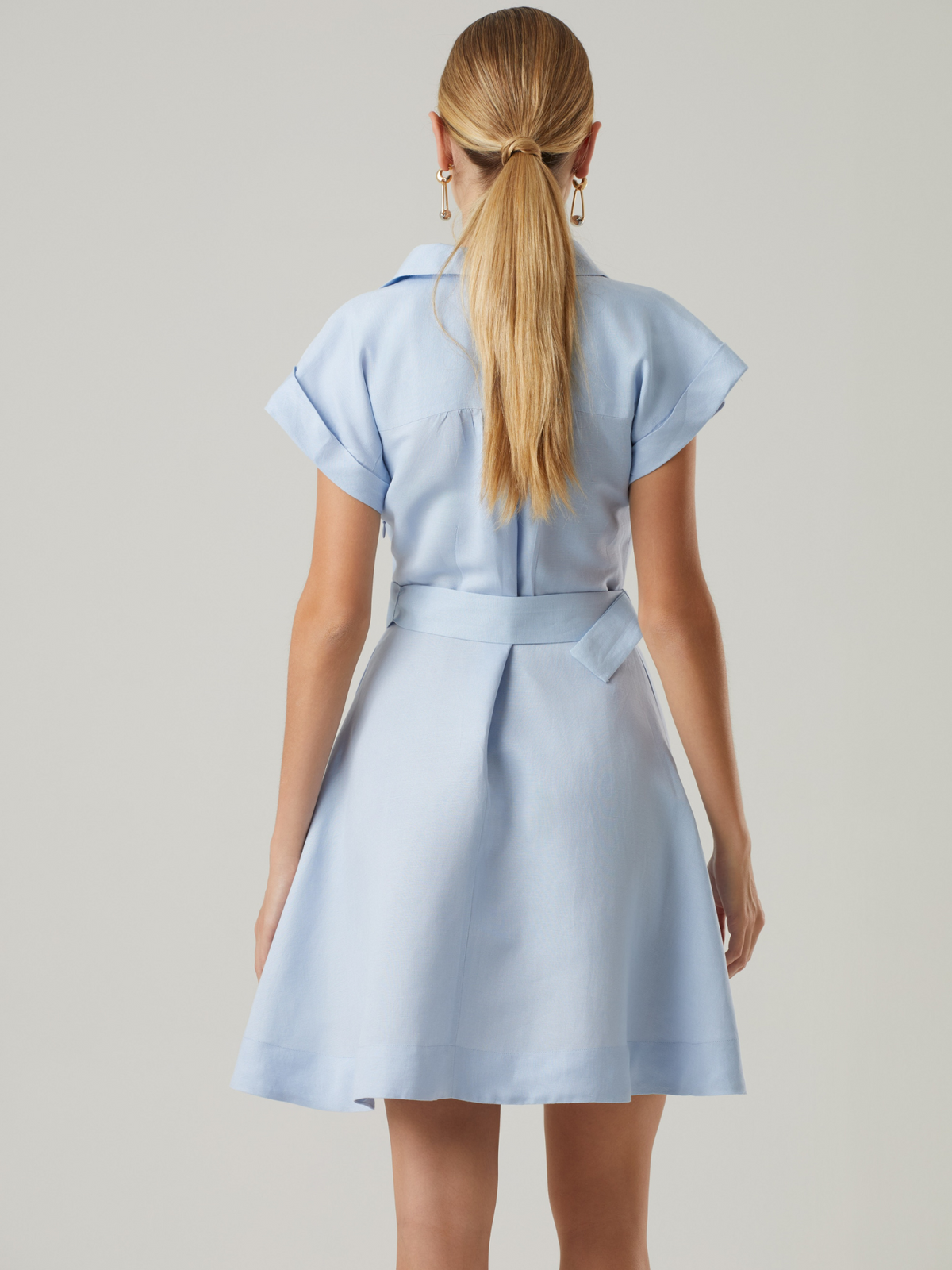 Este vestido corto combina un aire femenino con detalles clásicos. Tiene escote en V, botones en el frente y un cinturón con hebilla forrada que enmarca la cintura. Sus mangas cortas con pliegues y bolsillos decorativos en el pecho aportan un toque estructurado sin perder ligereza. Ideal para looks frescos y pulidos.