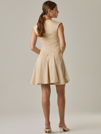 Vestido corto con silueta entallada en la parte superior y falda amplia. Tiene cuello cerrado tipo caja, mangas pequeñas que apenas cubren los hombros y costuras que definen la estructura del torso. La falda incorpora pliegues profundos que le dan volumen y movimiento. El largo llega por encima de la rodilla. Cierre invisible en la espalda.