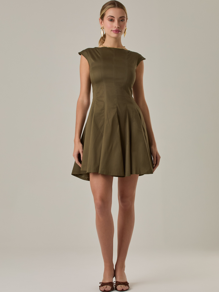 Vestido corto con silueta entallada en la parte superior y falda amplia. Tiene cuello cerrado tipo caja, mangas pequeñas que apenas cubren los hombros y costuras que definen la estructura del torso. La falda incorpora pliegues profundos que le dan volumen y movimiento. El largo llega por encima de la rodilla. Cierre invisible en la espalda.