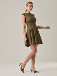 Vestido corto con silueta entallada en la parte superior y falda amplia. Tiene cuello cerrado tipo caja, mangas pequeñas que apenas cubren los hombros y costuras que definen la estructura del torso. La falda incorpora pliegues profundos que le dan volumen y movimiento. El largo llega por encima de la rodilla. Cierre invisible en la espalda.

Detalles:

Color: Olivo