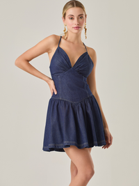 Es un vestido corto confeccionado en tela tipo denim de tono azul oscuro. Tiene escote en V pronunciado con tirantes delgados y ajustables. La parte superior se ajusta al cuerpo resaltando la cintura, mientras que la falda tiene un diseño con vuelo que aporta movimiento y feminidad. Es un vestido casual pero elegante, ideal para días cálidos o eventos informales.