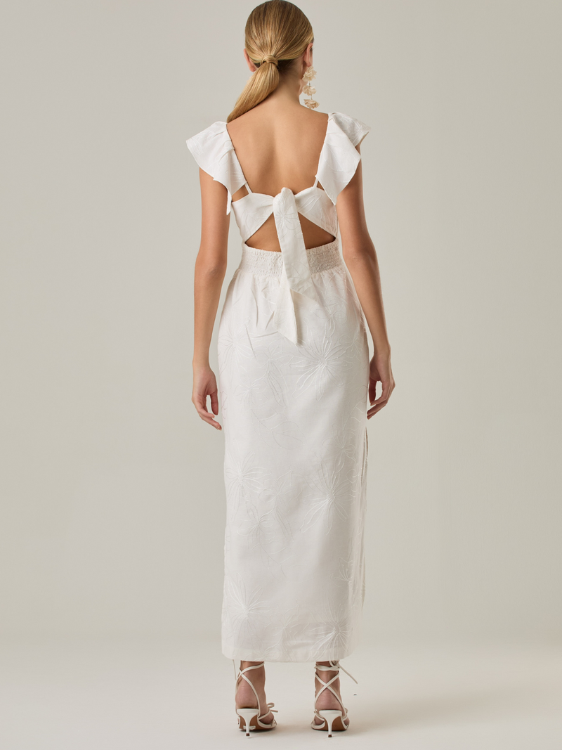 Este vestido tiene un aire delicado y femenino. Es de largo midi, con escote en “V” profundo al frente y en la espalda, y detalles de volantes en los tirantes. La parte trasera se ajusta con un lazo, dejando una abertura que resalta la cintura. El textil tiene un sutil bordado de hojas y flores, aportando textura sin ser recargado. El corte favorece la silueta y la abertura lateral en la falda añade movimiento al caminar. Ideal para eventos elegantes en climas cálidos.