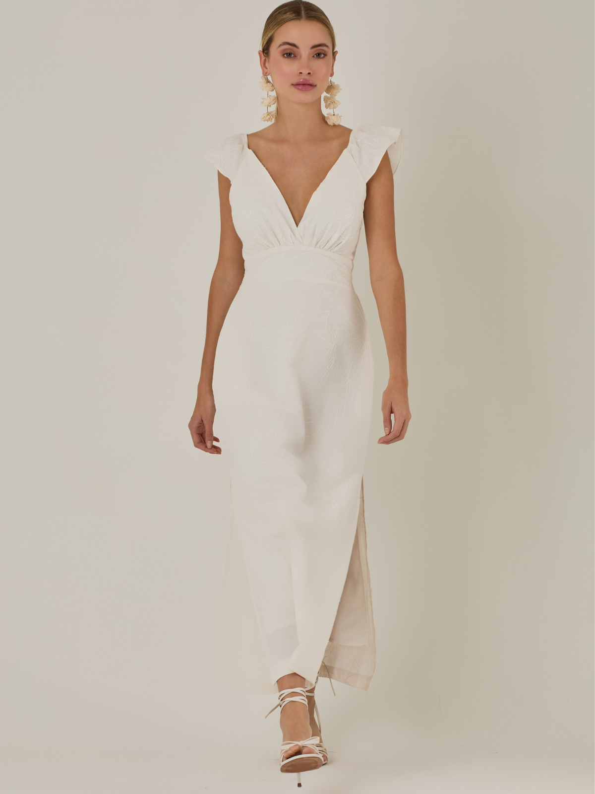 Este vestido tiene un aire delicado y femenino. Es de largo midi, con escote en “V” profundo al frente y en la espalda, y detalles de volantes en los tirantes. La parte trasera se ajusta con un lazo, dejando una abertura que resalta la cintura. El textil tiene un sutil bordado de hojas y flores, aportando textura sin ser recargado. El corte favorece la silueta y la abertura lateral en la falda añade movimiento al caminar. Ideal para eventos elegantes en climas cálidos.