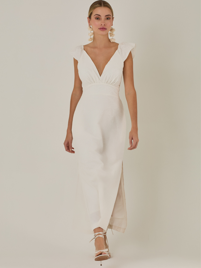 Este vestido tiene un aire delicado y femenino. Es de largo midi, con escote en “V” profundo al frente y en la espalda, y detalles de volantes en los tirantes. La parte trasera se ajusta con un lazo, dejando una abertura que resalta la cintura. El textil tiene un sutil bordado de hojas y flores, aportando textura sin ser recargado. El corte favorece la silueta y la abertura lateral en la falda añade movimiento al caminar. Ideal para eventos elegantes en climas cálidos.