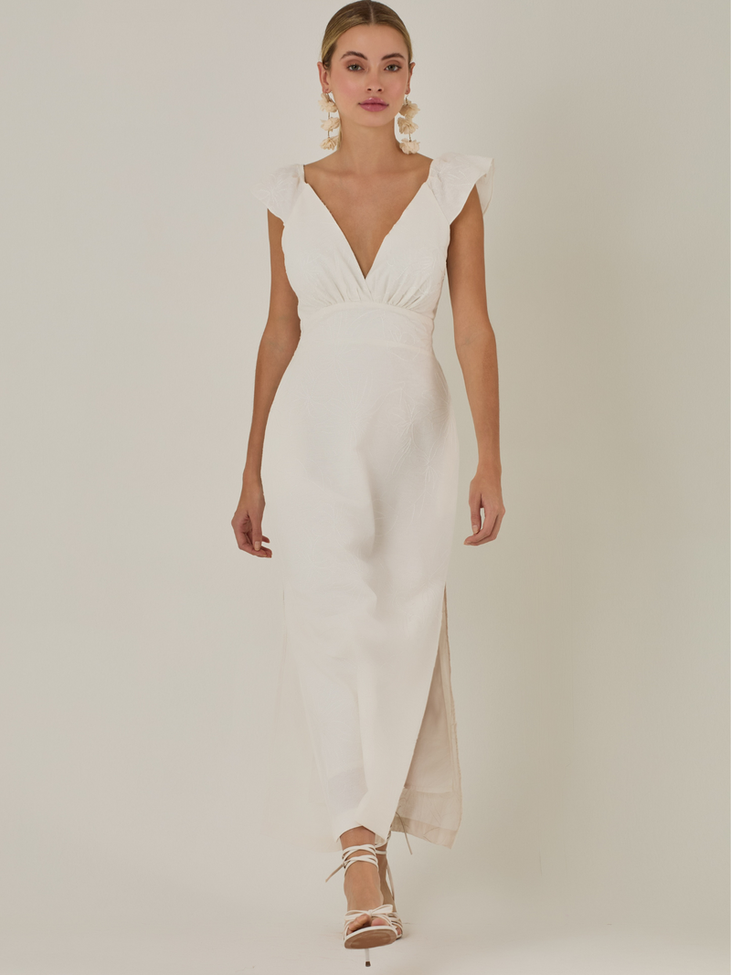 Este vestido tiene un aire delicado y femenino. Es de largo midi, con escote en “V” profundo al frente y en la espalda, y detalles de volantes en los tirantes. La parte trasera se ajusta con un lazo, dejando una abertura que resalta la cintura. El textil tiene un sutil bordado de hojas y flores, aportando textura sin ser recargado. El corte favorece la silueta y la abertura lateral en la falda añade movimiento al caminar. Ideal para eventos elegantes en climas cálidos.