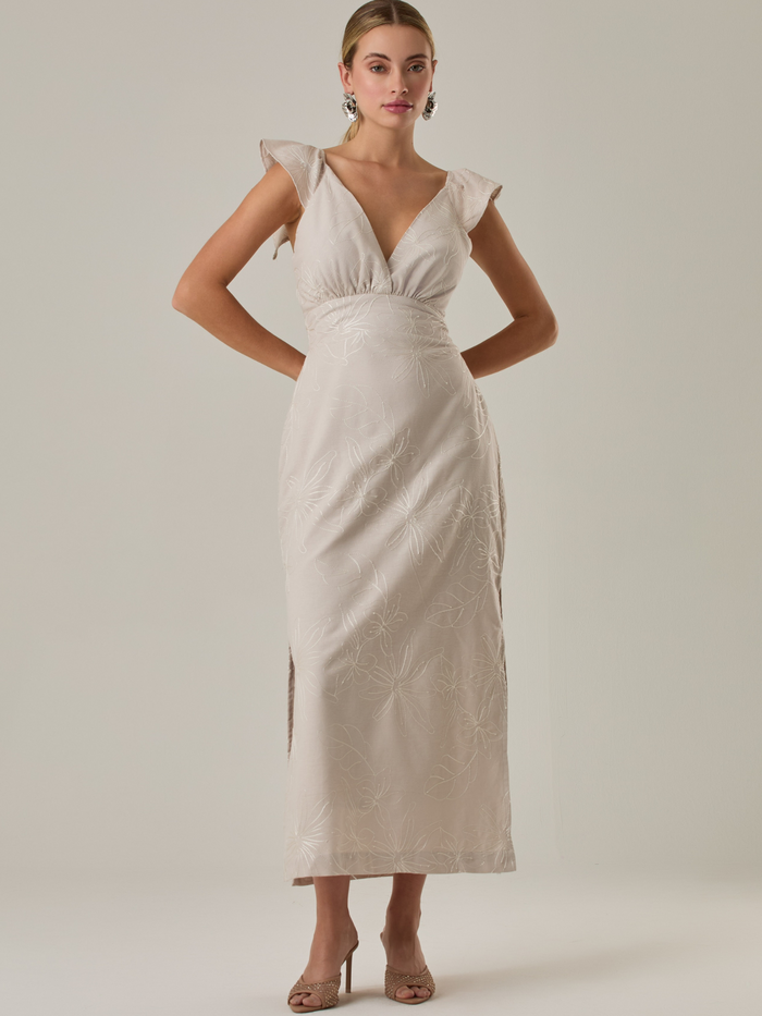 Este vestido tiene un aire delicado y femenino. Es de largo midi, con escote en “V” profundo al frente y en la espalda, y detalles de volantes en los tirantes. La parte trasera se ajusta con un lazo, dejando una abertura que resalta la cintura. El textil tiene un sutil bordado de hojas y flores, aportando textura sin ser recargado. El corte favorece la silueta y la abertura lateral en la falda añade movimiento al caminar.