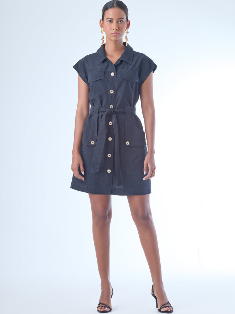 Vestido corto de estilo camisero con botones grandes al frente. Cuenta con cuello clásico, mangas cortas tipo casquillo y bolsillos de parche en el pecho y la parte baja del frente, que aportan un aire utilitario. La cintura se ajusta con un cinturón del mismo tejido, creando una silueta definida. Su diseño combina funcionalidad y sofisticación de manera equilibrada.

Detalles:

Color: Negro