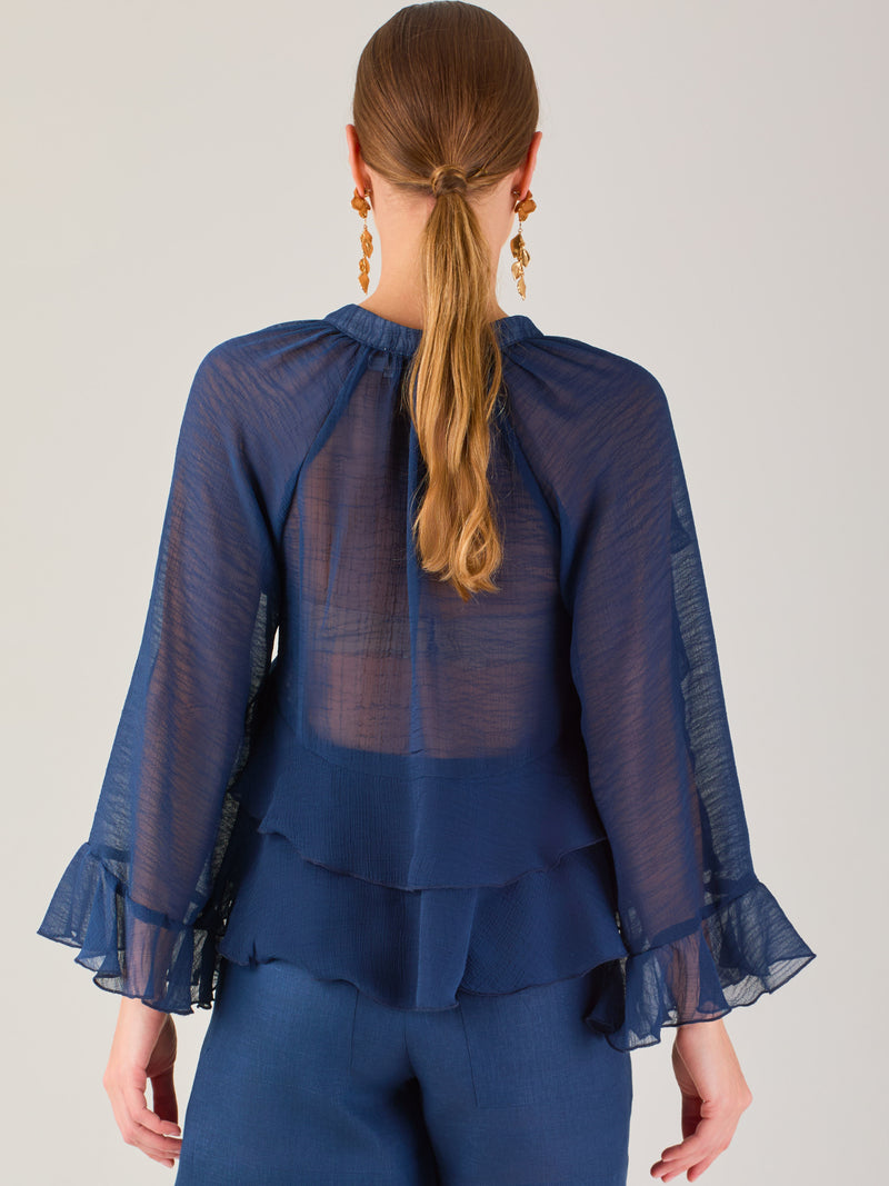 Ligera y romántica, esta blusa de textura vaporosa destaca por sus volantes en capas que aportan movimiento y fluidez. El escote en V y las mangas amplias con acabado ondulado añaden un toque delicado y sofisticado. Ideal para ocasiones especiales, cenas o estilismos casuales con un aire femenino y refinado.