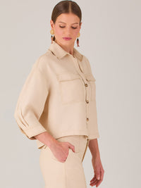 Esta blusa presenta un diseño tipo chaqueta corta (cropped) en un tono beige neutro, ideal para un look sofisticado y contemporáneo. Está confeccionada en una tela estructurada que mantiene su forma, aportando un aire pulido al conjunto. Tiene cuello camisero clásico, cierre frontal con botones metálicos visibles, y dos bolsillos de parche en el pecho con solapa y botón decorativo, que refuerzan el estilo utilitario.