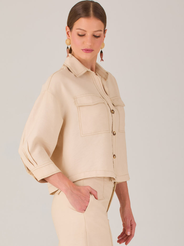 Esta blusa presenta un diseño tipo chaqueta corta (cropped) en un tono beige neutro, ideal para un look sofisticado y contemporáneo. Está confeccionada en una tela estructurada que mantiene su forma, aportando un aire pulido al conjunto. Tiene cuello camisero clásico, cierre frontal con botones metálicos visibles, y dos bolsillos de parche en el pecho con solapa y botón decorativo, que refuerzan el estilo utilitario.