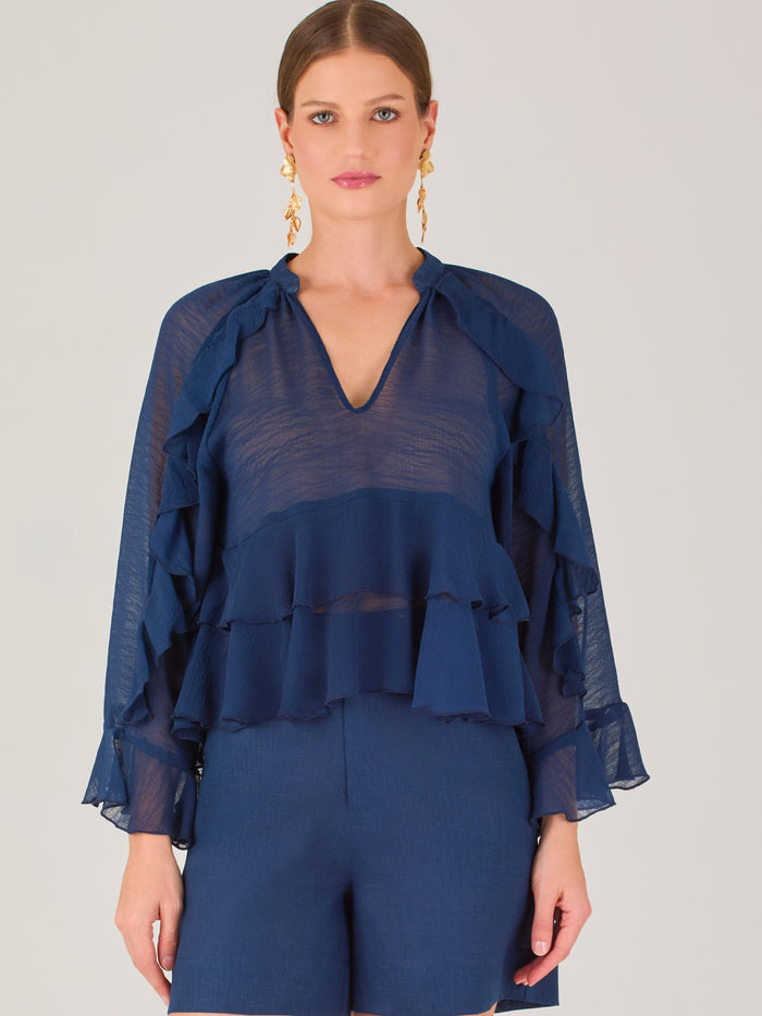 Ligera y romántica, esta blusa de textura vaporosa destaca por sus volantes en capas que aportan movimiento y fluidez. El escote en V y las mangas amplias con acabado ondulado añaden un toque delicado y sofisticado. Ideal para ocasiones especiales, cenas o estilismos casuales con un aire femenino y refinado.