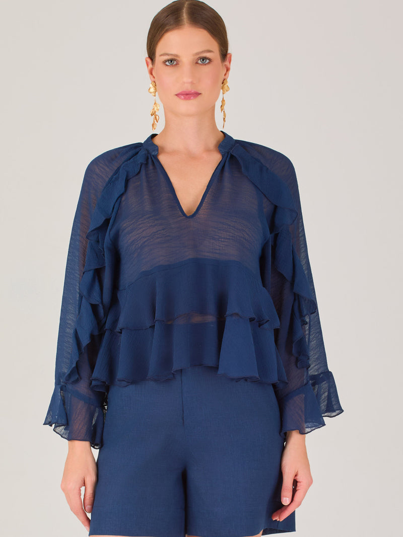 Ligera y romántica, esta blusa de textura vaporosa destaca por sus volantes en capas que aportan movimiento y fluidez. El escote en V y las mangas amplias con acabado ondulado añaden un toque delicado y sofisticado. Ideal para ocasiones especiales, cenas o estilismos casuales con un aire femenino y refinado.