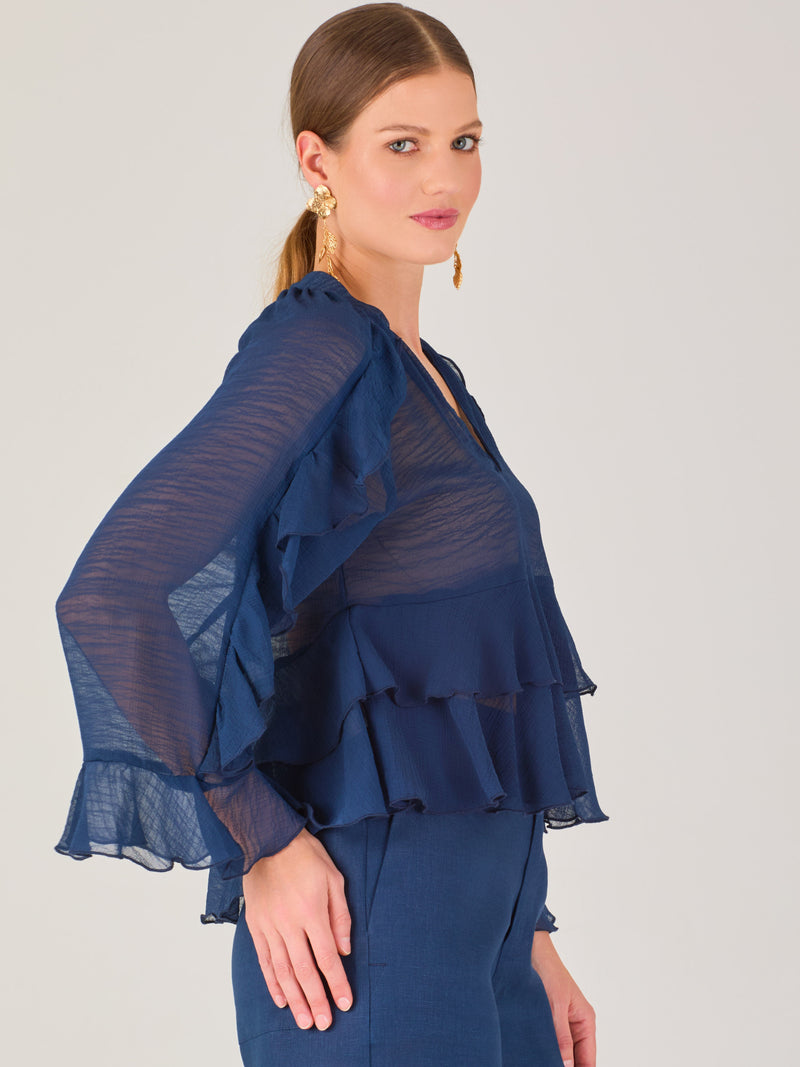 Ligera y romántica, esta blusa de textura vaporosa destaca por sus volantes en capas que aportan movimiento y fluidez. El escote en V y las mangas amplias con acabado ondulado añaden un toque delicado y sofisticado. Ideal para ocasiones especiales, cenas o estilismos casuales con un aire femenino y refinado.