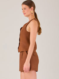Esta blusa tipo chaleco es ideal para climas cálidos o para crear looks sofisticados con un aire casual. Presenta un corte recto ligeramente entallado que acentúa la silueta de forma sutil, y un escote en “V” que estiliza el cuello. Cuenta con una botonadura frontal compuesta por cinco botones oscuros que contrastan elegantemente con el tono neutro de la prenda. También incorpora bolsillos falsos decorativos a ambos lados del frente, que añaden un detalle sutil al diseño minimalista. Esta pieza es versátil 