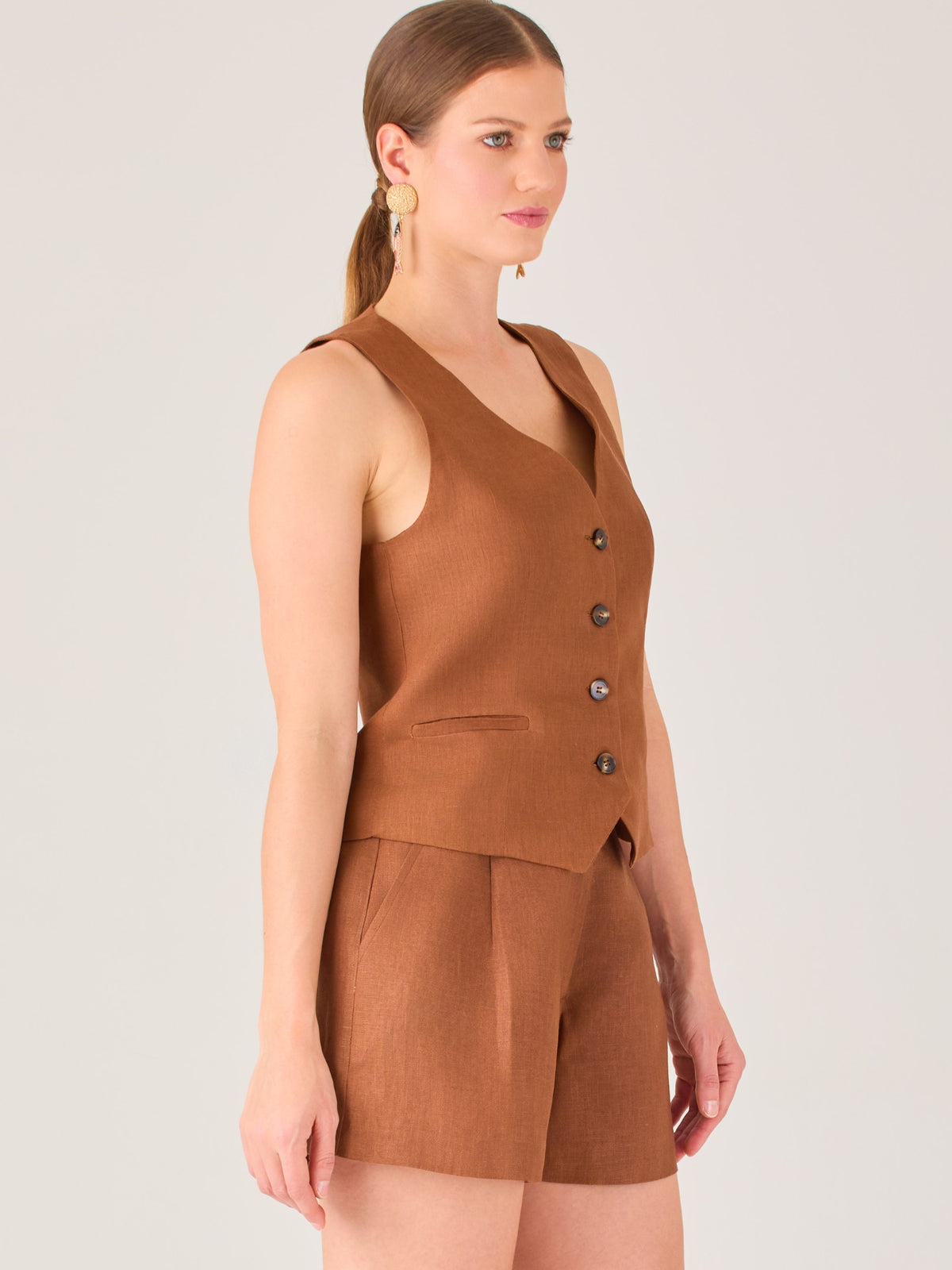 Esta blusa tipo chaleco es ideal para climas cálidos o para crear looks sofisticados con un aire casual. Presenta un corte recto ligeramente entallado que acentúa la silueta de forma sutil, y un escote en “V” que estiliza el cuello. Cuenta con una botonadura frontal compuesta por cinco botones oscuros que contrastan elegantemente con el tono neutro de la prenda. También incorpora bolsillos falsos decorativos a ambos lados del frente, que añaden un detalle sutil al diseño minimalista. Esta pieza es versátil 