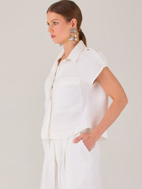 Blusa blanca de estilo camisero con un diseño moderno y minimalista. Cuenta con cuello clásico, tapeta oculta para los botones y mangas cortas tipo caída con detalle de charreteras en los hombros. Posee dos bolsillos de parche en el pecho que le aportan un toque utilitario y estructurado. Su corte recto y fluido la hace ideal tanto para looks casuales como elegantes
