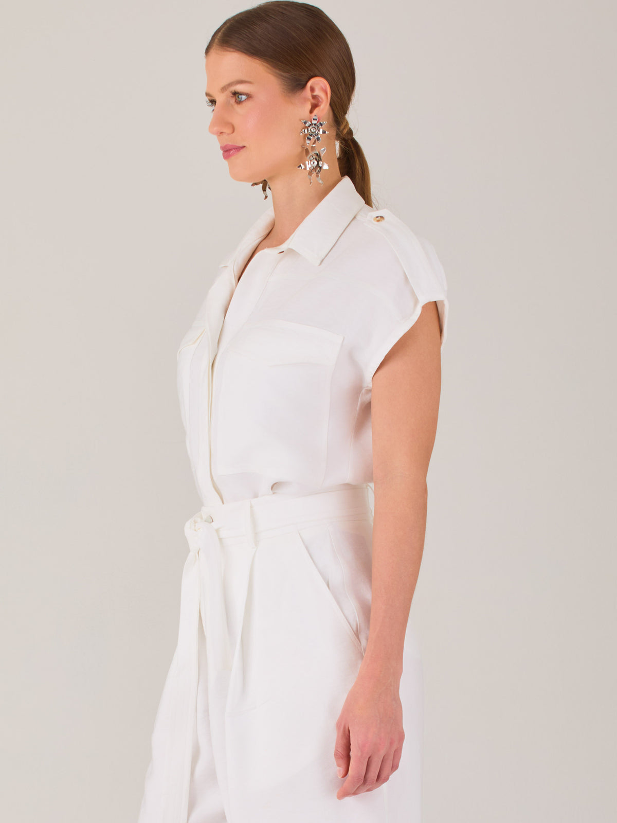 Blusa blanca de estilo camisero con un diseño moderno y minimalista. Cuenta con cuello clásico, tapeta oculta para los botones y mangas cortas tipo caída con detalle de charreteras en los hombros. Posee dos bolsillos de parche en el pecho que le aportan un toque utilitario y estructurado. Su corte recto y fluido la hace ideal tanto para looks casuales como elegantes