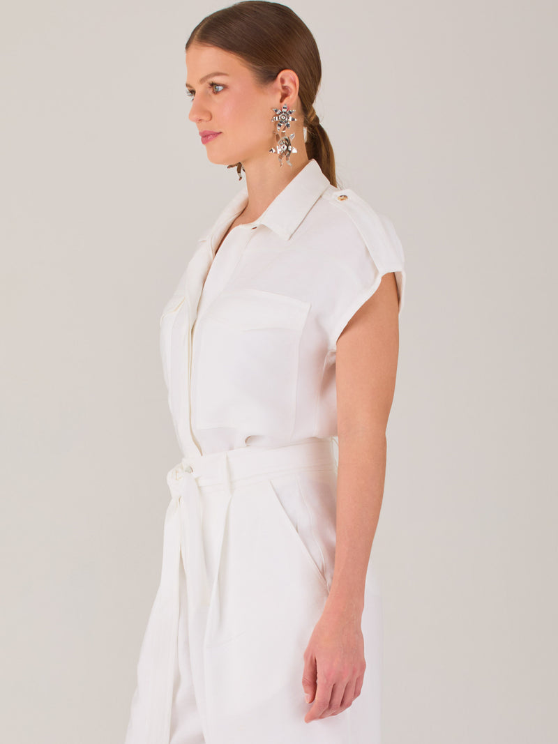 Blusa blanca de estilo camisero con un diseño moderno y minimalista. Cuenta con cuello clásico, tapeta oculta para los botones y mangas cortas tipo caída con detalle de charreteras en los hombros. Posee dos bolsillos de parche en el pecho que le aportan un toque utilitario y estructurado. Su corte recto y fluido la hace ideal tanto para looks casuales como elegantes