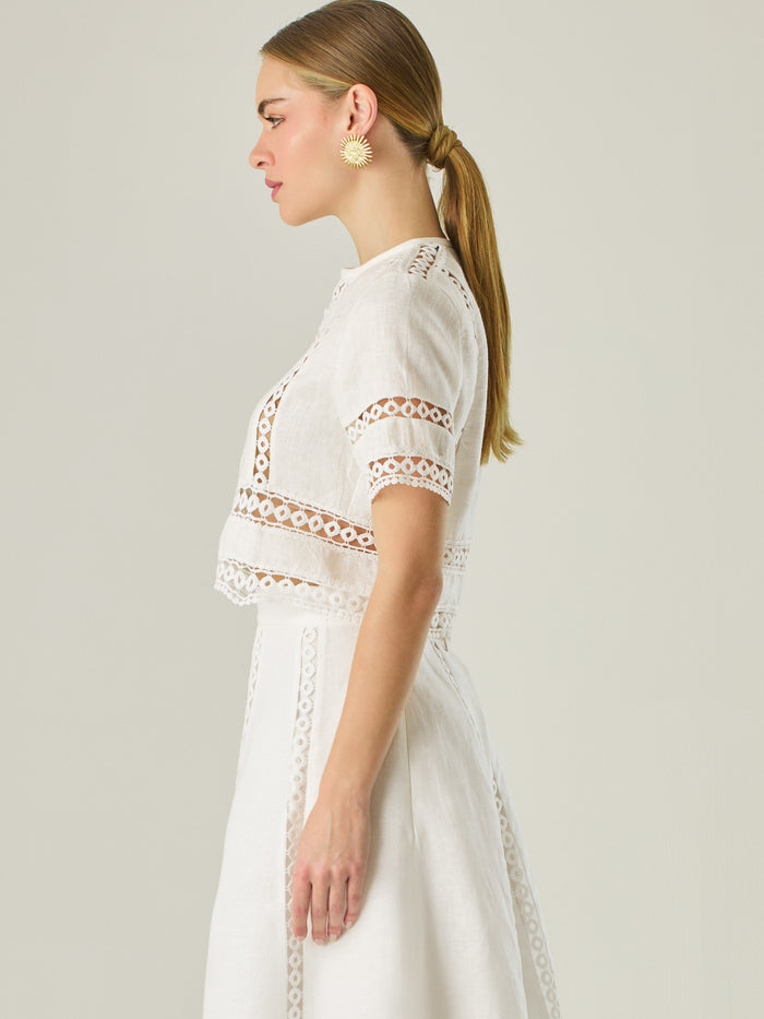 Esta elegante blusa blanca está confeccionada en un tejido liviano tipo lino, ideal para climas cálidos. Presenta un corte recto y estructurado, con mangas cortas y cuello cerrado redondo. Lo más distintivo son sus detalles decorativos en encaje tipo guipiur dispuesto en franjas verticales y horizontales sobre el frente, mangas y parte inferior, que crean un efecto visual sofisticado y femenino al mostrar sutilmente la piel, su diseño combina frescura y refinamiento, siendo perfecta para looks veraniegos co