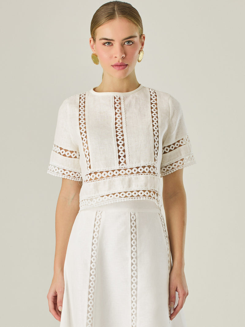 Esta elegante blusa blanca está confeccionada en un tejido liviano tipo lino, ideal para climas cálidos. Presenta un corte recto y estructurado, con mangas cortas y cuello cerrado redondo. Lo más distintivo son sus detalles decorativos en encaje tipo guipiur dispuesto en franjas verticales y horizontales sobre el frente, mangas y parte inferior, que crean un efecto visual sofisticado y femenino al mostrar sutilmente la piel, su diseño combina frescura y refinamiento, siendo perfecta para looks veraniegos co