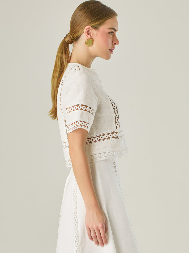 Esta elegante blusa blanca está confeccionada en un tejido liviano tipo lino, ideal para climas cálidos. Presenta un corte recto y estructurado, con mangas cortas y cuello cerrado redondo. Lo más distintivo son sus detalles decorativos en encaje tipo guipiur dispuesto en franjas verticales y horizontales sobre el frente, mangas y parte inferior, que crean un efecto visual sofisticado y femenino al mostrar sutilmente la piel, su diseño combina frescura y refinamiento, siendo perfecta para looks veraniegos co